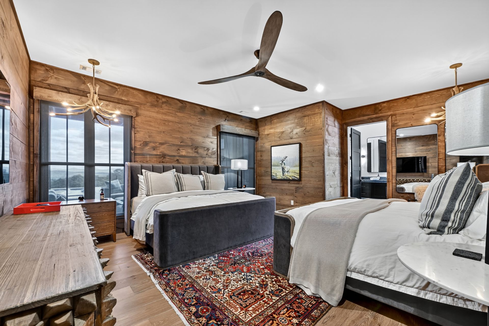Bedroom with two beds, wooden walls, a ceiling fan, and a view through a large window.