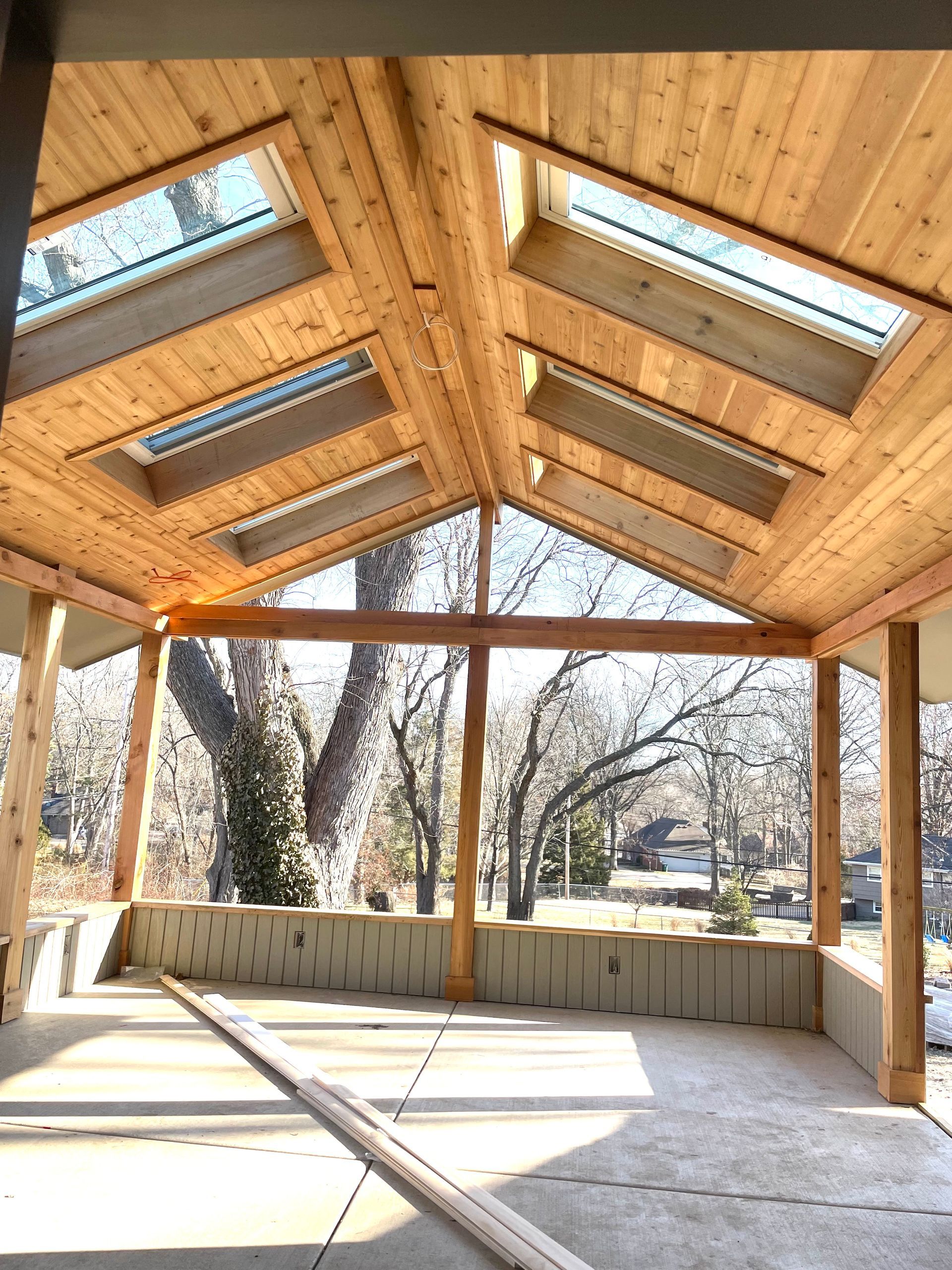 Open-air wooden pavilion with skylights and a concrete floor in a park.