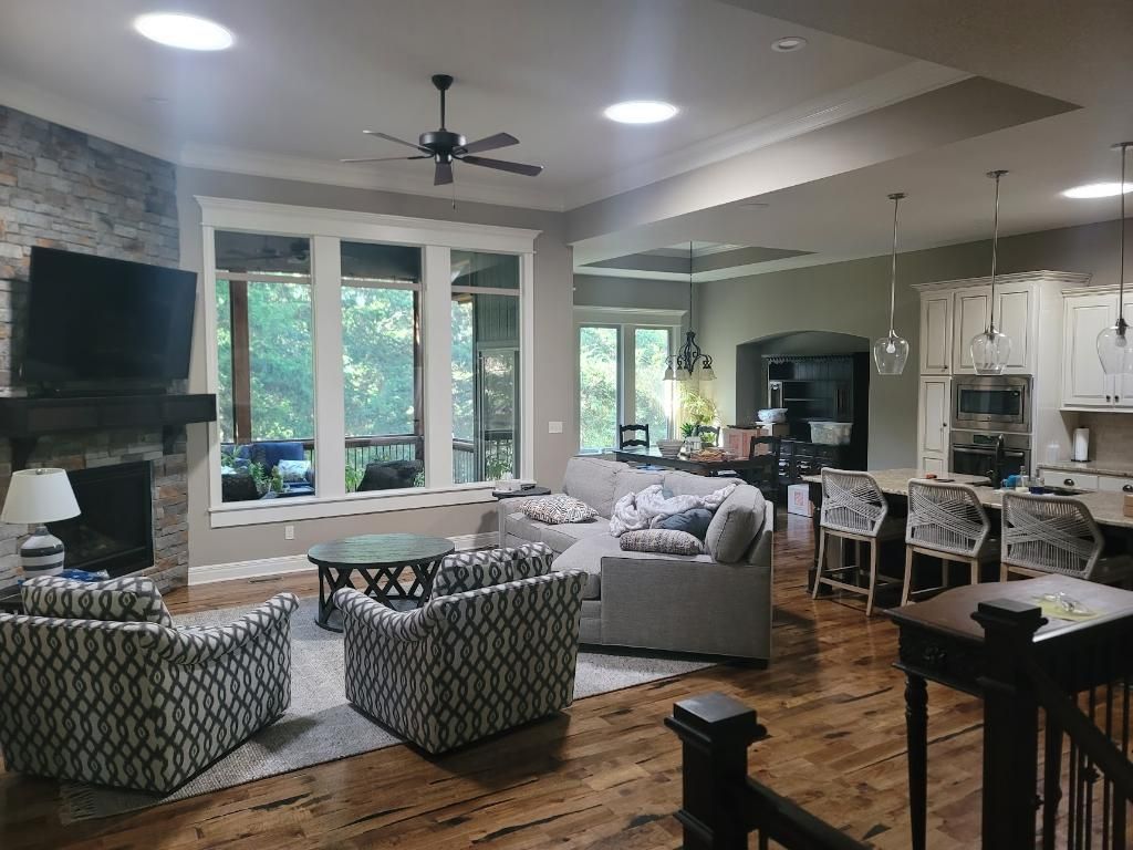 Bright open-concept living room with fireplace, gray sofa, patterned chairs, kitchen bar stools, and large windows