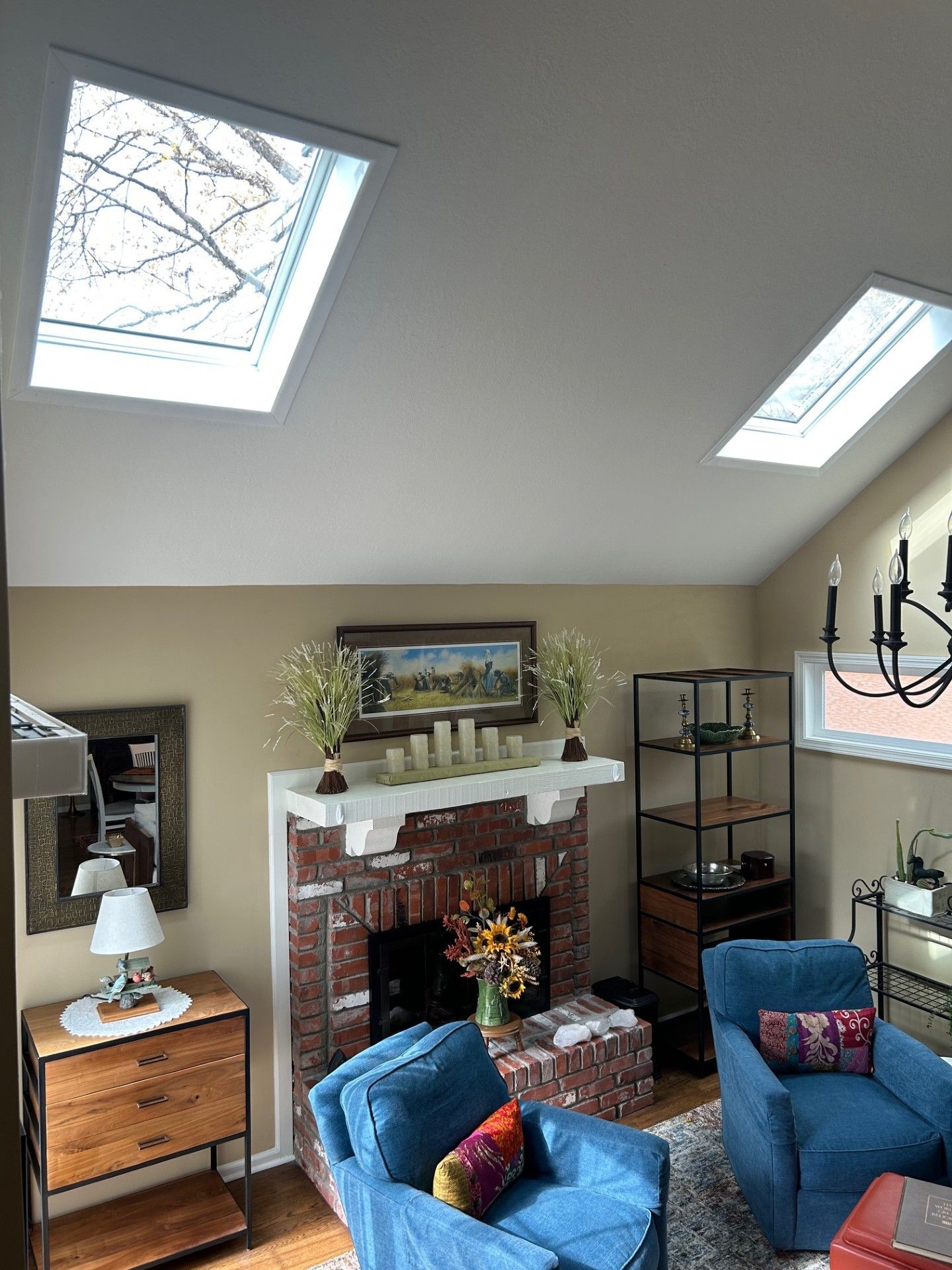 Bright living room with skylights, white brick fireplace, blue chairs, and a glass shelving unit.