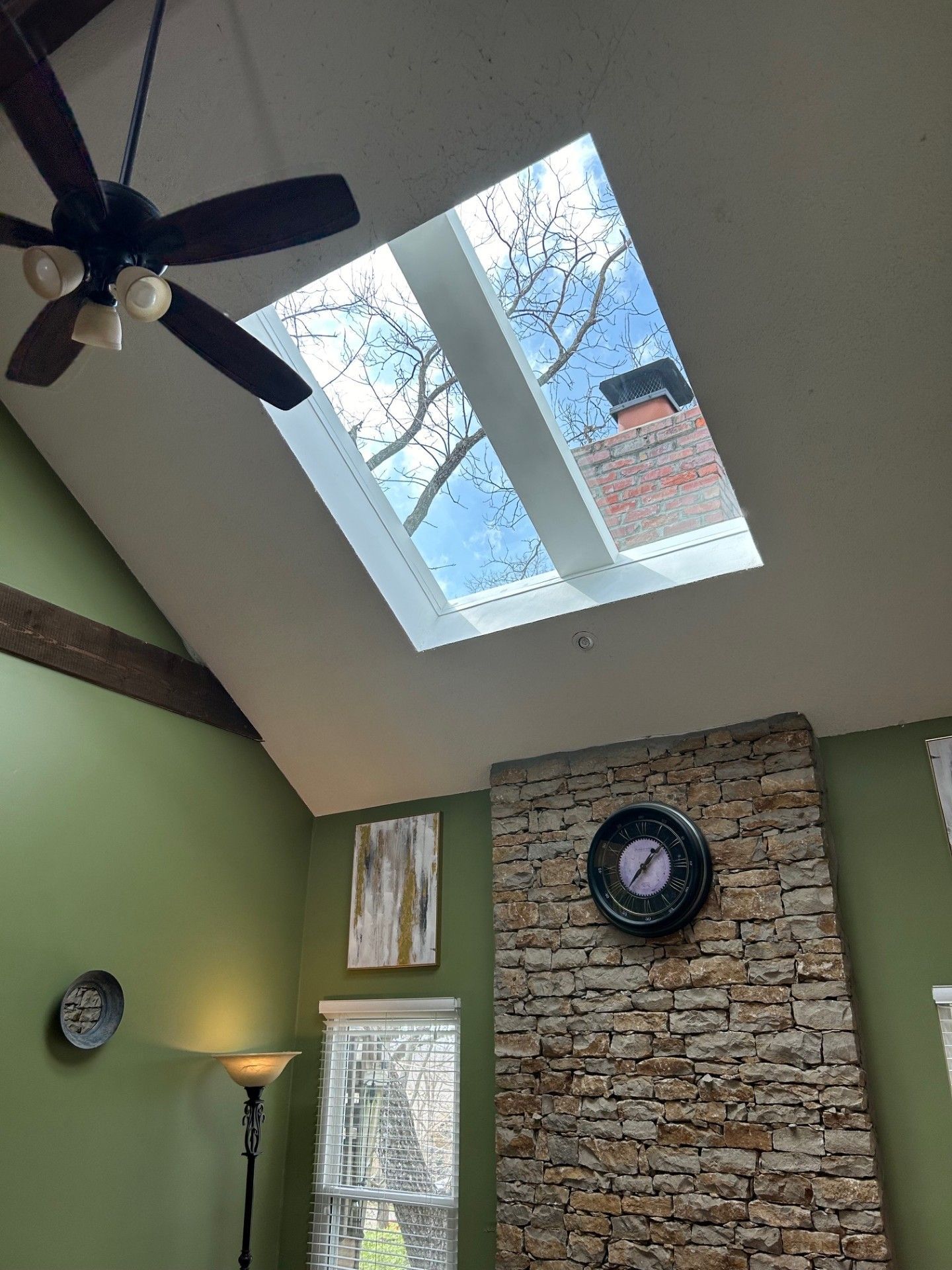Living room with vaulted ceiling, skylight, ceiling fan, stone accent wall, clock, and lamp