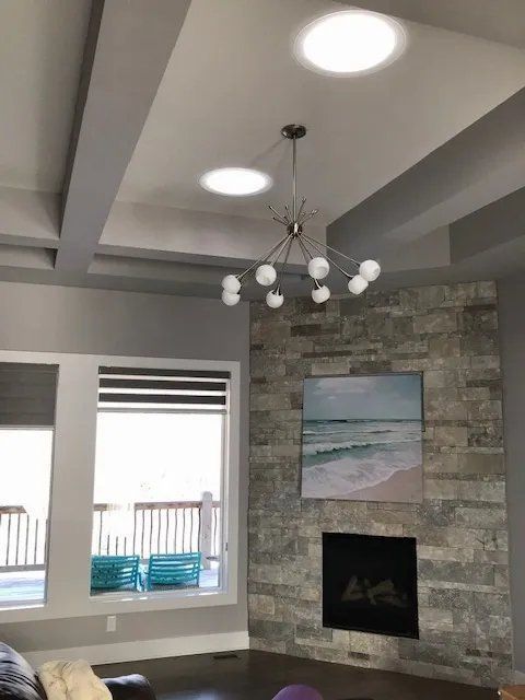 Modern living room with coffered ceiling, stone fireplace, beach painting, and windows overlooking a balcony.