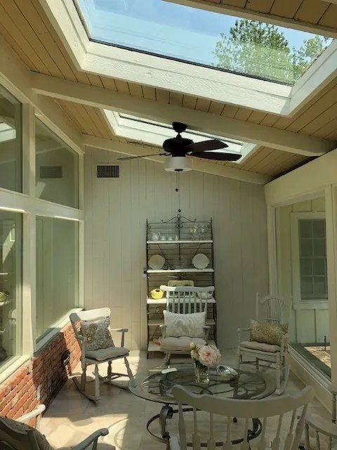 Covered patio with skylights, ceiling fan, round table, chairs, and shelving against white walls.
