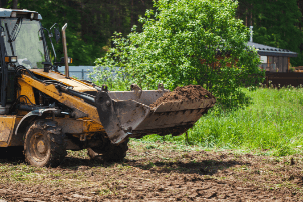 Yellow backhoe with dirt in bucket, clearing a field. Green foliage and small building in the background.
