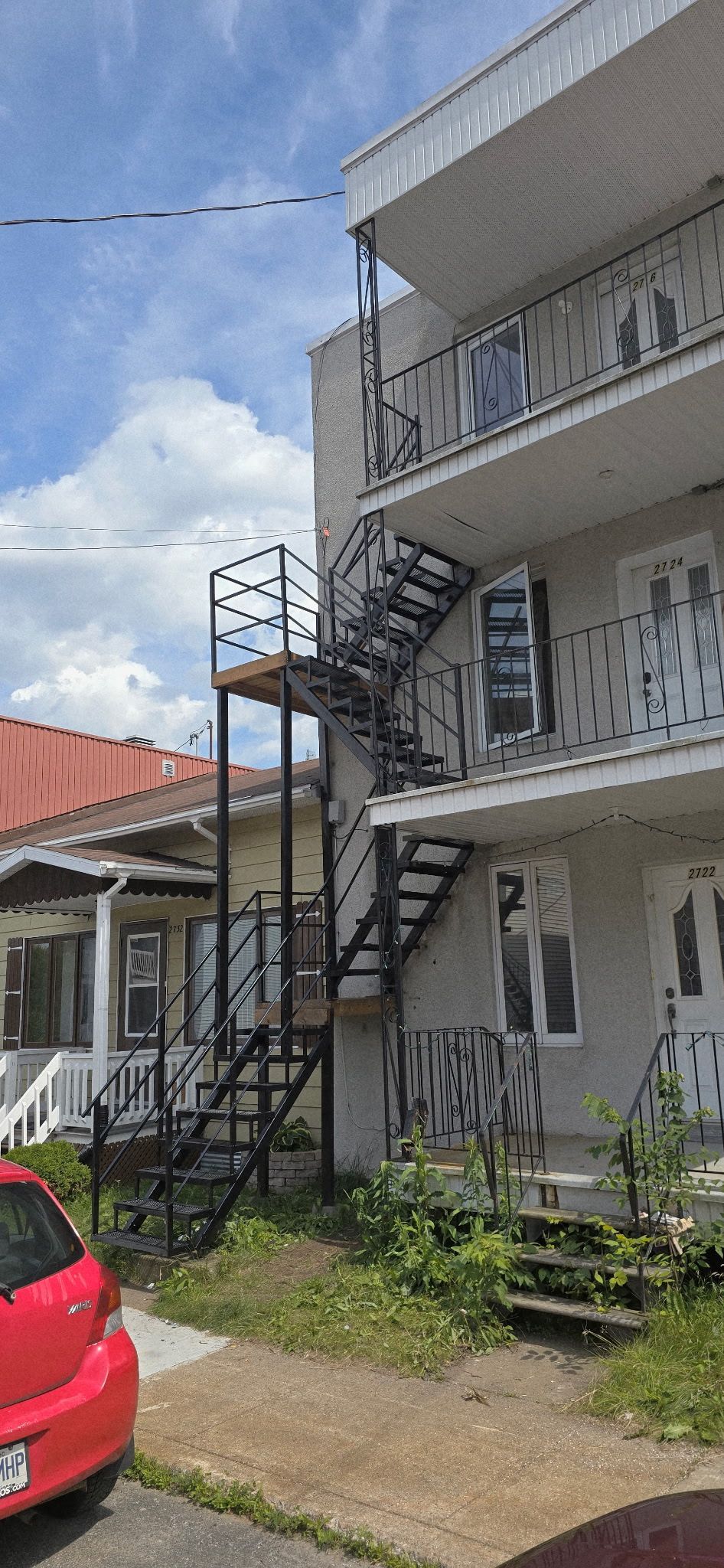 Une voiture rouge garée devant un immeuble à plusieurs étages avec un escalier de secours en métal.