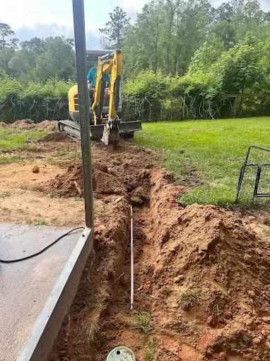 Excavator digging a trench in a grassy yard; next to a patio. Sunny day.
