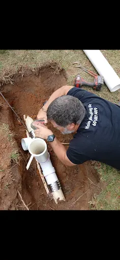 A plumber working on PVC pipes in a dirt trench. The man wears a black shirt and uses a tool.