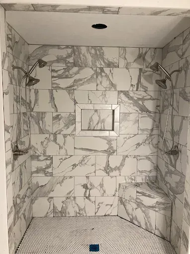 Marble-tiled shower with two showerheads, a recessed shelf, and a small bench.