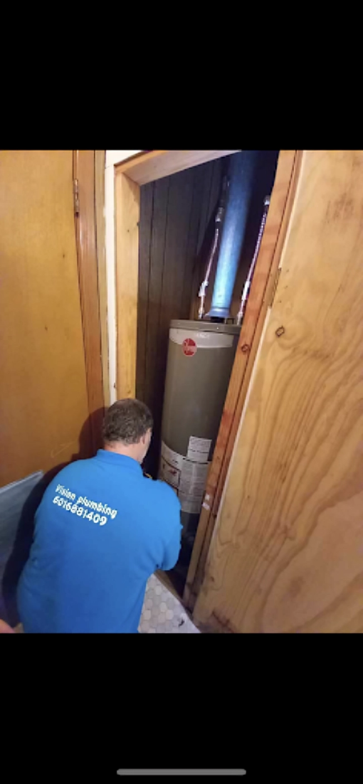 A plumber in a blue shirt inspects a hot water heater inside a closet.