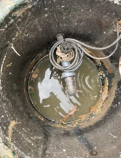 Inside a dark tank, a submersible pump floats in murky water. Gray cable coils above.