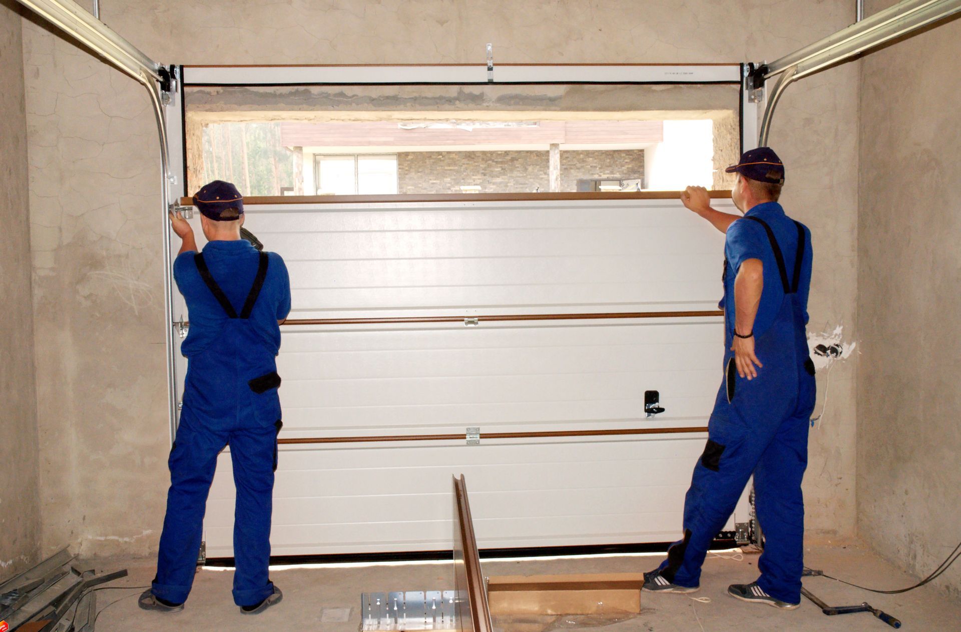 Two workers in blue overalls install a large white garage door in a construction site