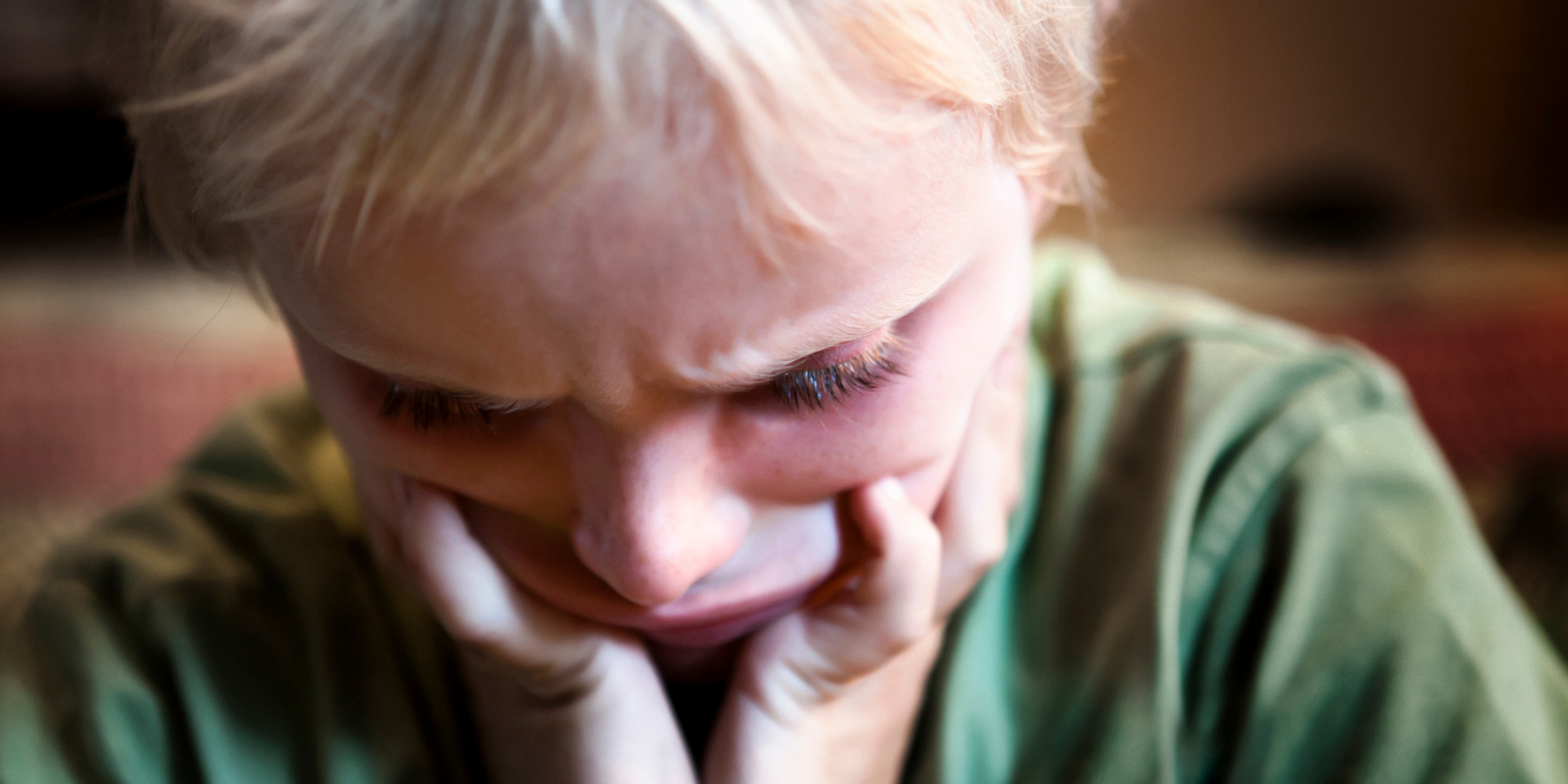Crying child with head down and hands on cheeks, wearing a green shirt indoors