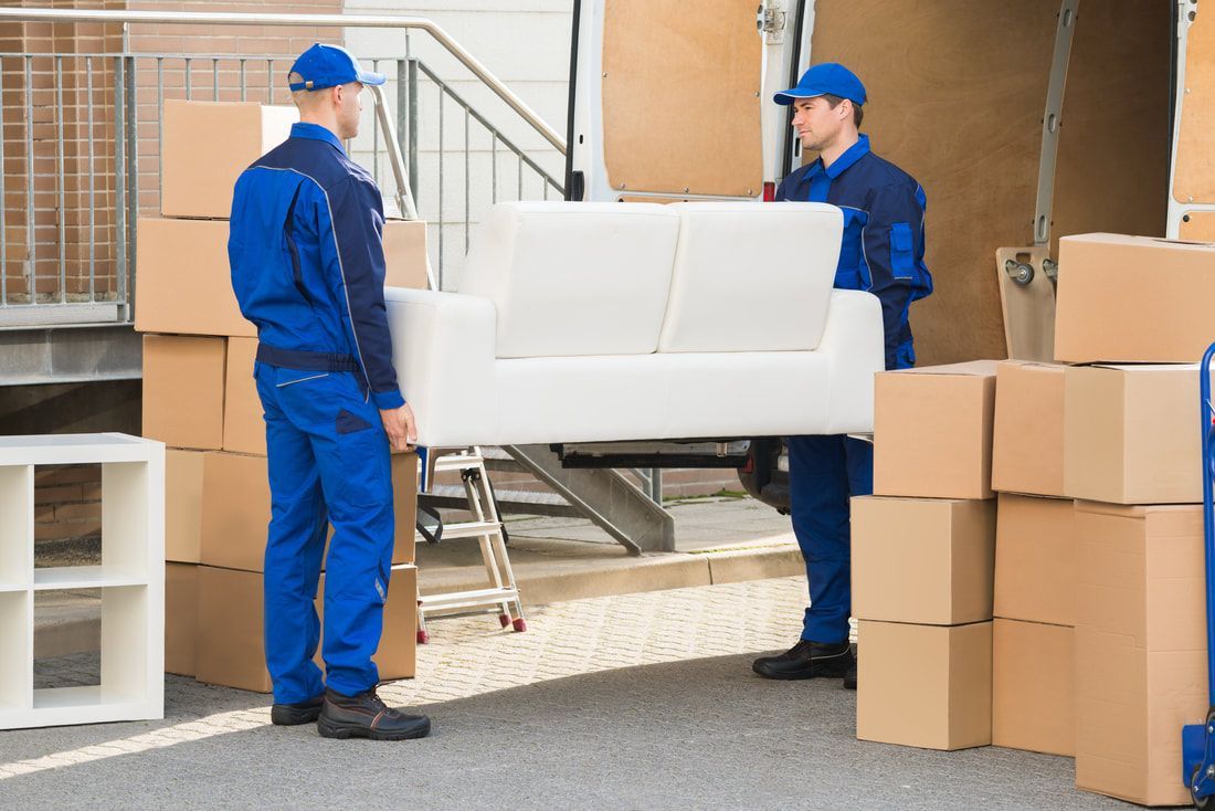 Two movers in blue uniforms carrying a white sofa towards a truck; moving boxes around.