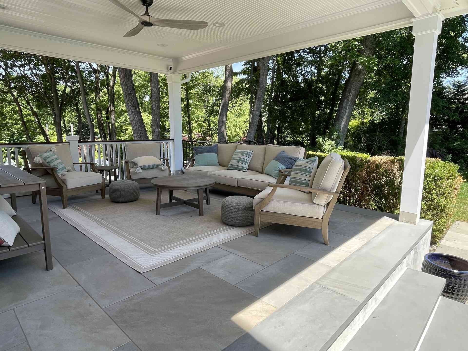 Covered patio with cushioned seating, coffee table, and rug overlooking a wooded yard