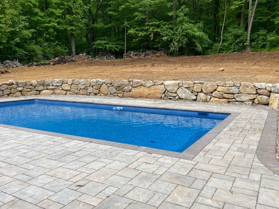 Blue swimming pool beside stone wall and paved patio, surrounded by green trees