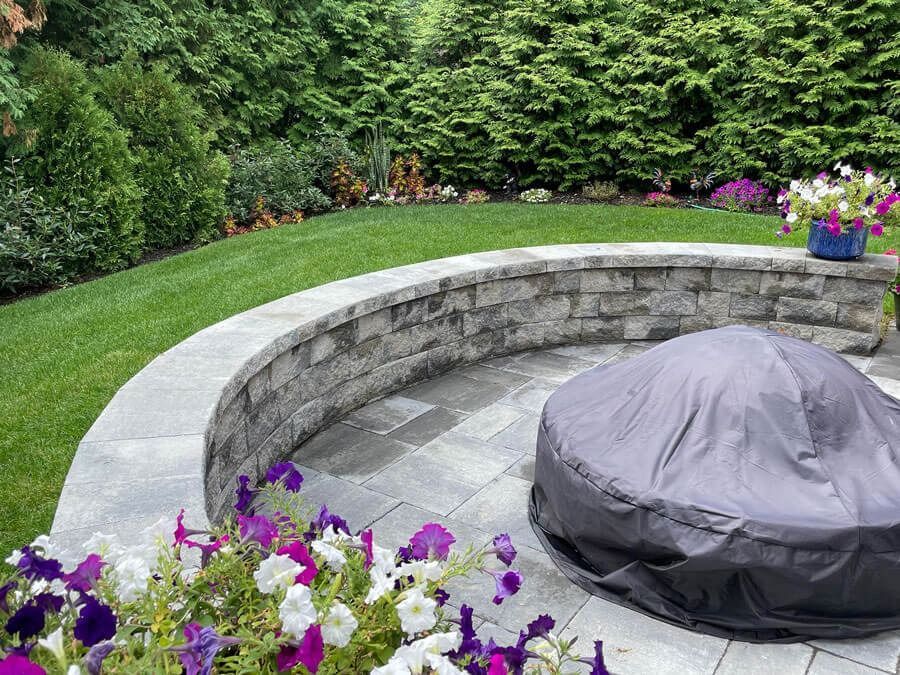 Stone fire pit in a landscaped garden with a curved bench, green lawn, and purple flowers
