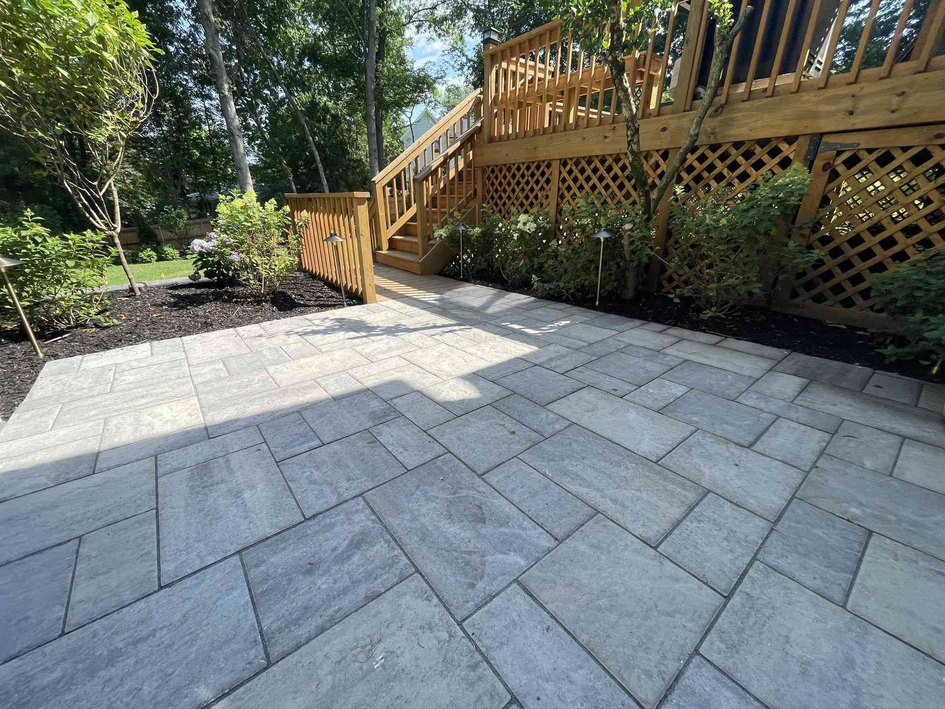 Stone patio with a wooden deck and stairs beside a landscaped garden in bright sunlight