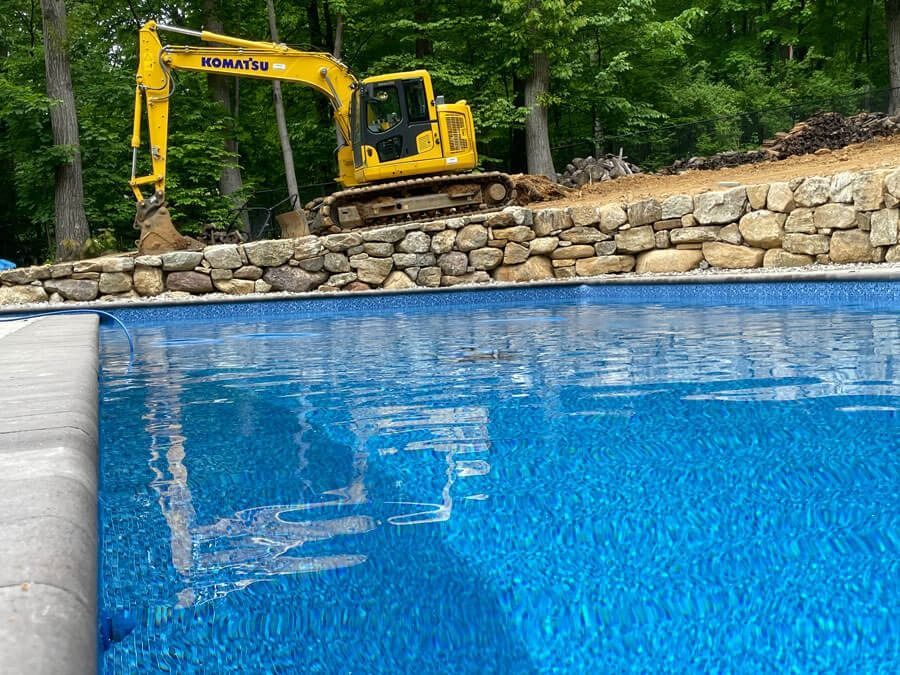 Yellow excavator behind stone wall beside a bright blue pool in a wooded yard