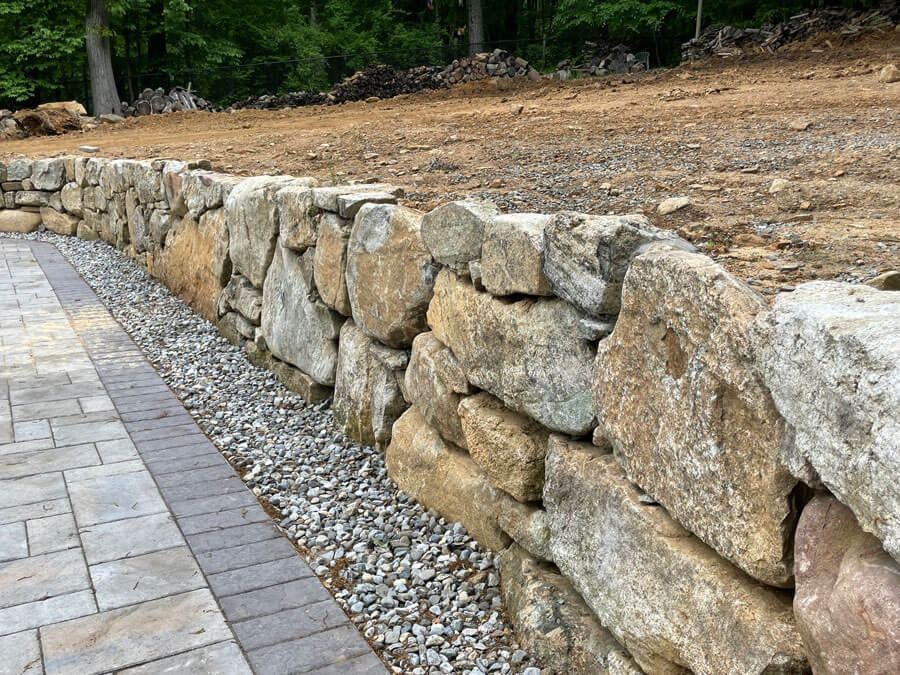 Stone retaining wall beside a paver walkway along a gravel landscaped slope with trees in the background