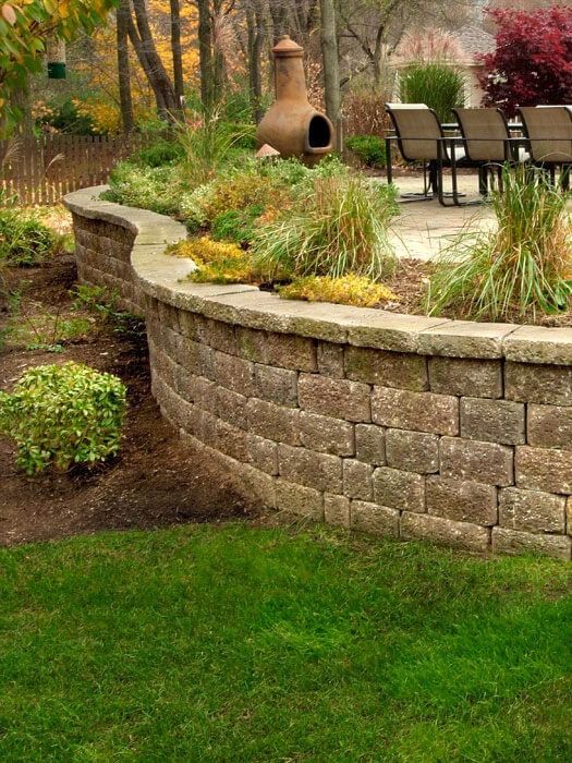 Curved stone garden wall with shrubs, patio chairs, and a clay chiminea in a landscaped yard