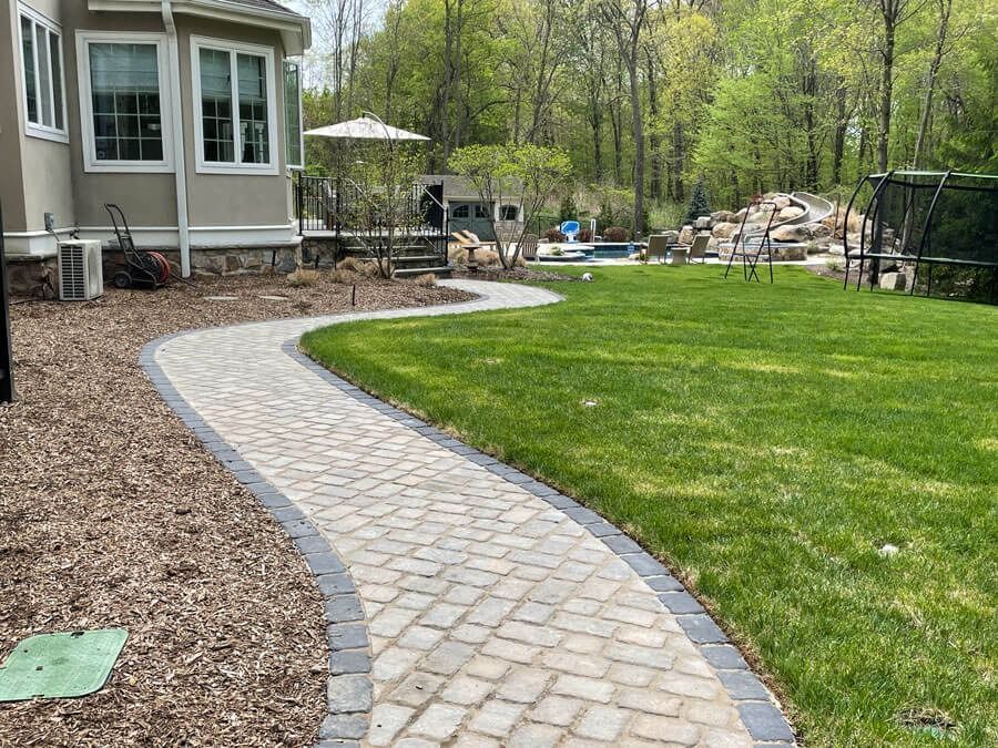 Curving stone path beside a house leads through a green backyard with trees and a patio.
