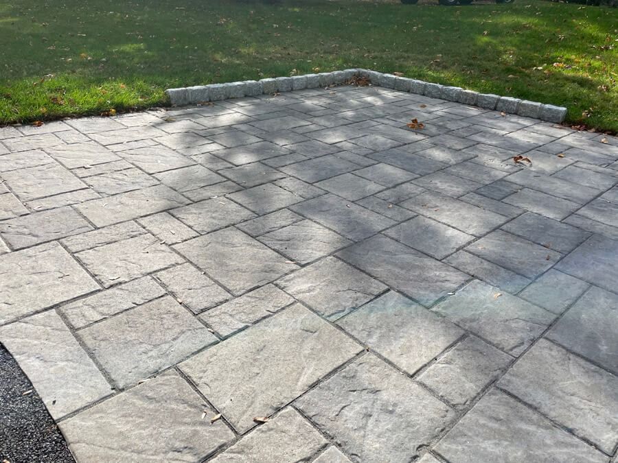 Empty stone patio with a raised curb, bordered by grass and dappled tree shadows