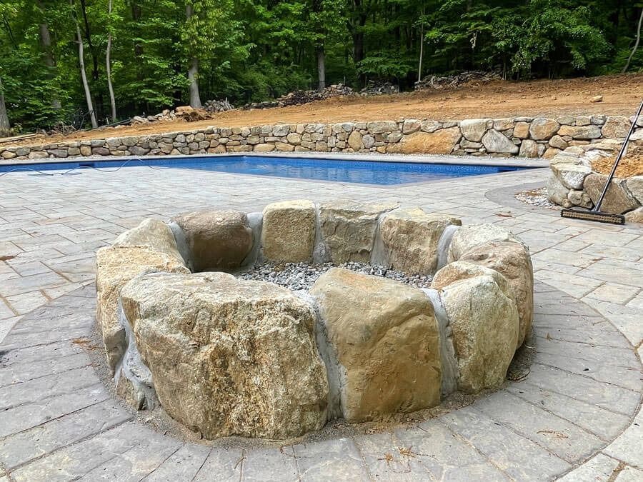 Stone fire pit beside a paved pool deck with trees and a stone retaining wall in the background