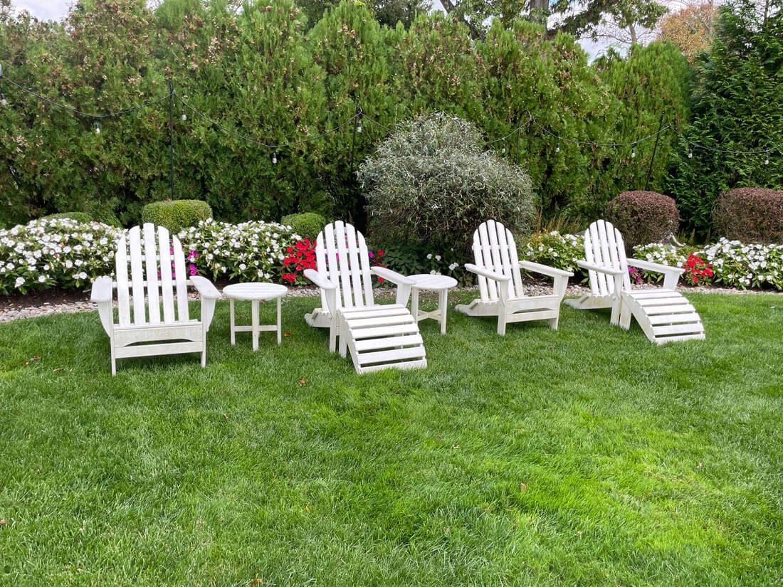 White Adirondack chairs on a green lawn in a garden, with bushes and flowers in the background.