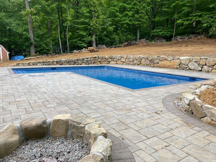Backyard pool beside a stone patio, with trees and a stone retaining wall in the background