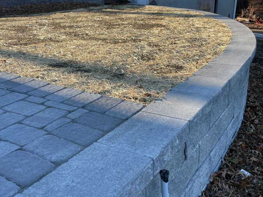 Raised stone planter with dry grass beside a paved walkway and curved retaining wall