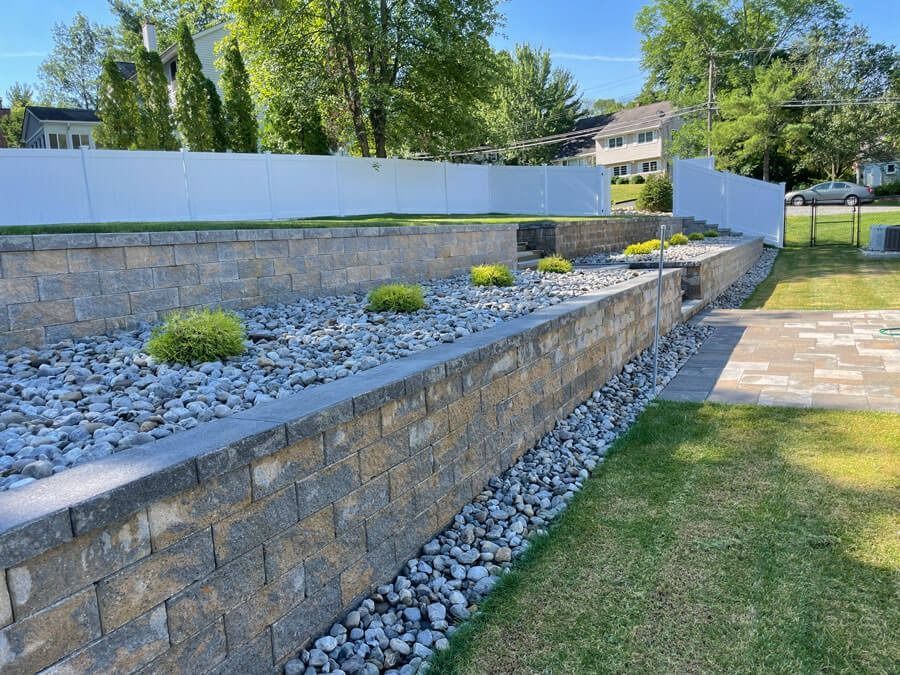 Stone retaining wall with white fence and landscaped rocks beside a grassy yard under trees