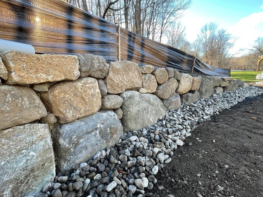 Stone retaining wall with gravel base along a roadside under bare trees