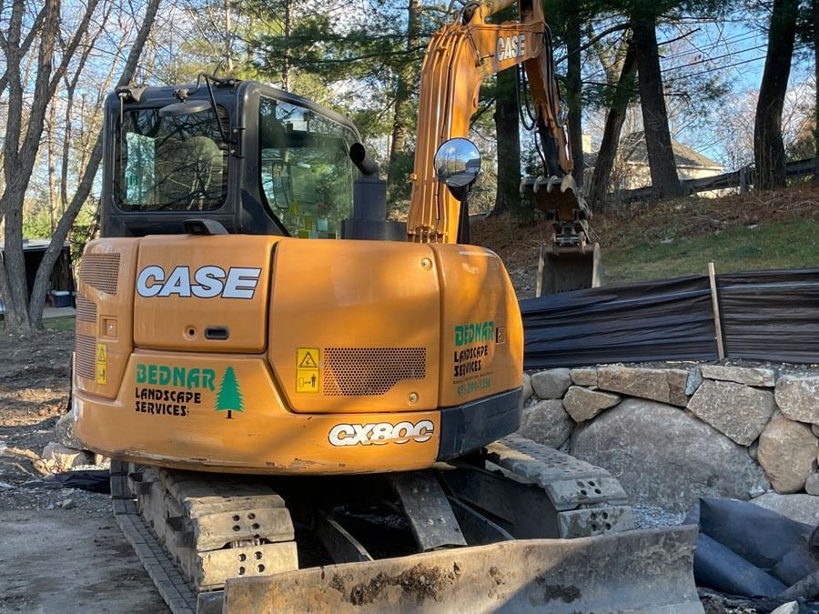 Orange excavator parked on a foundation site beside stacked stones and black plastic sheeting.