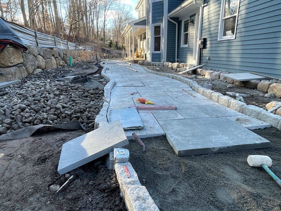 Partially finished concrete walkway beside a blue house, with construction materials and rubble along the path