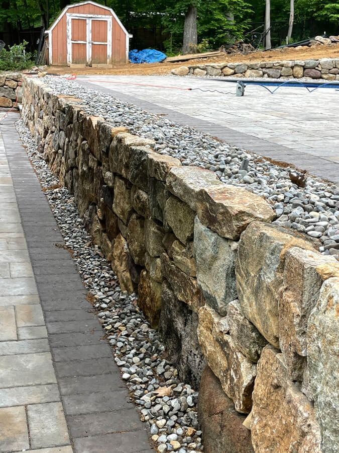 Stone retaining wall beside a paved path, with a small brown shed and gravel area in the background