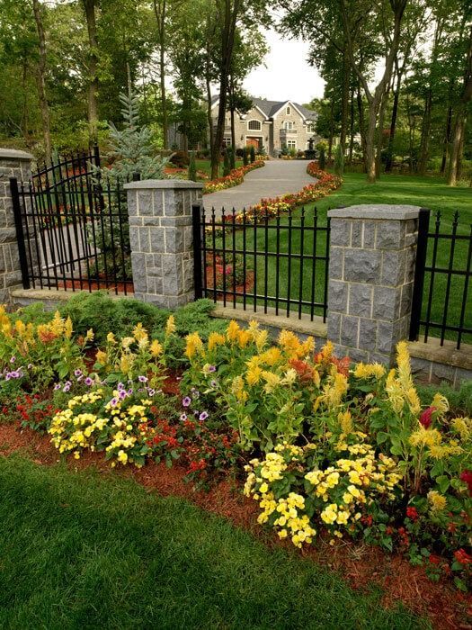 Front gate with stone pillars and black iron fence beside a colorful flower bed leading to a house