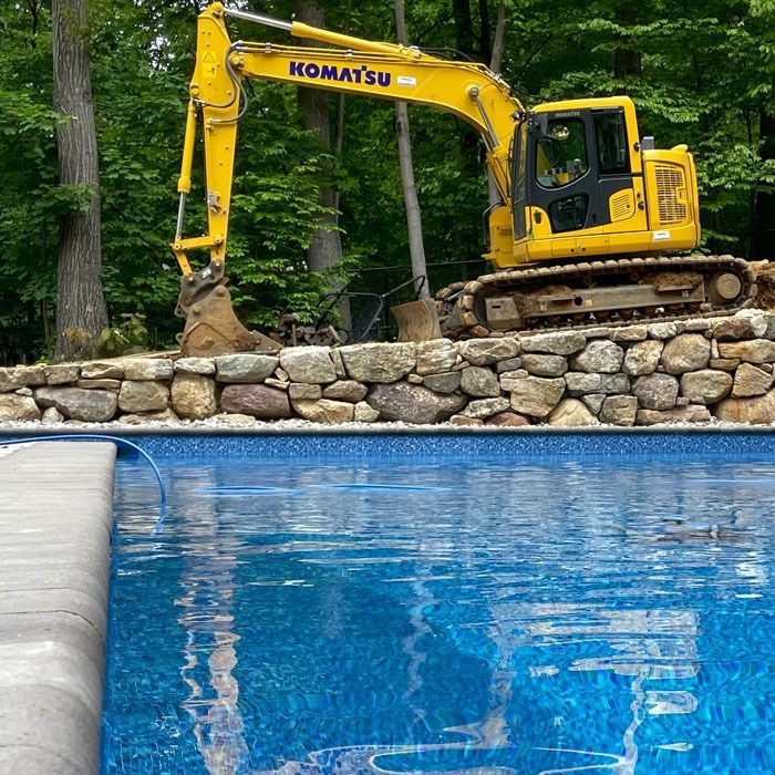 Yellow excavator on a stone wall beside a blue pool, digging near a wooded area.