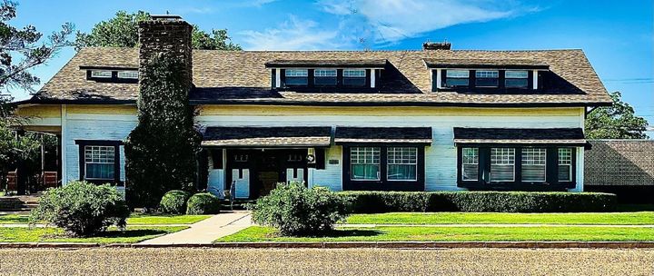 A weathered, one-story white house with a stone chimney, dark-trimmed windows, and dormers under a blue sky.