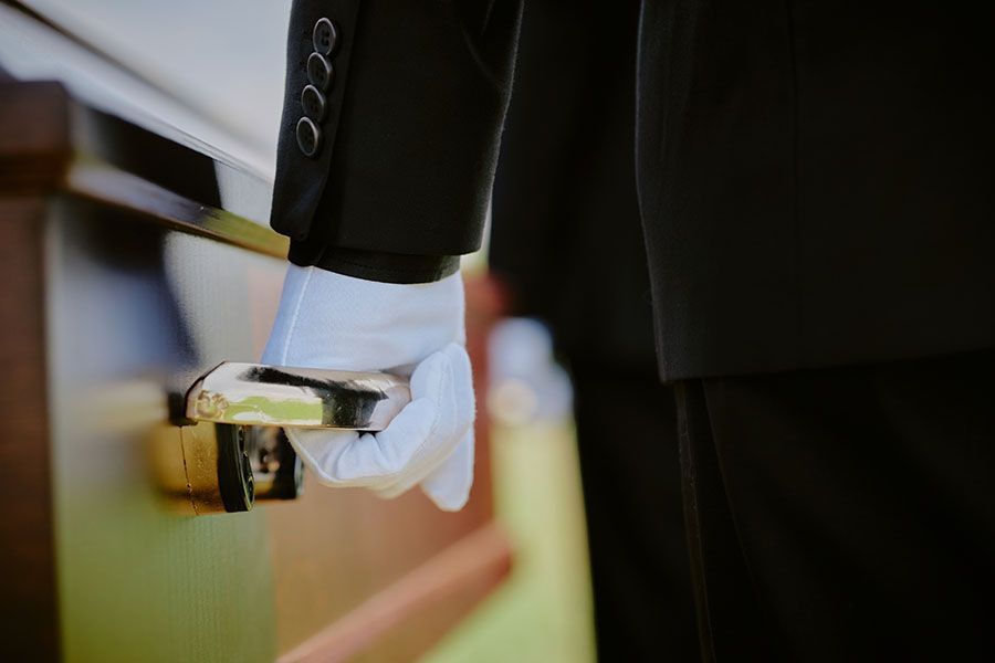 A person in a black suit and white gloves holds the handle of a dark wood casket.