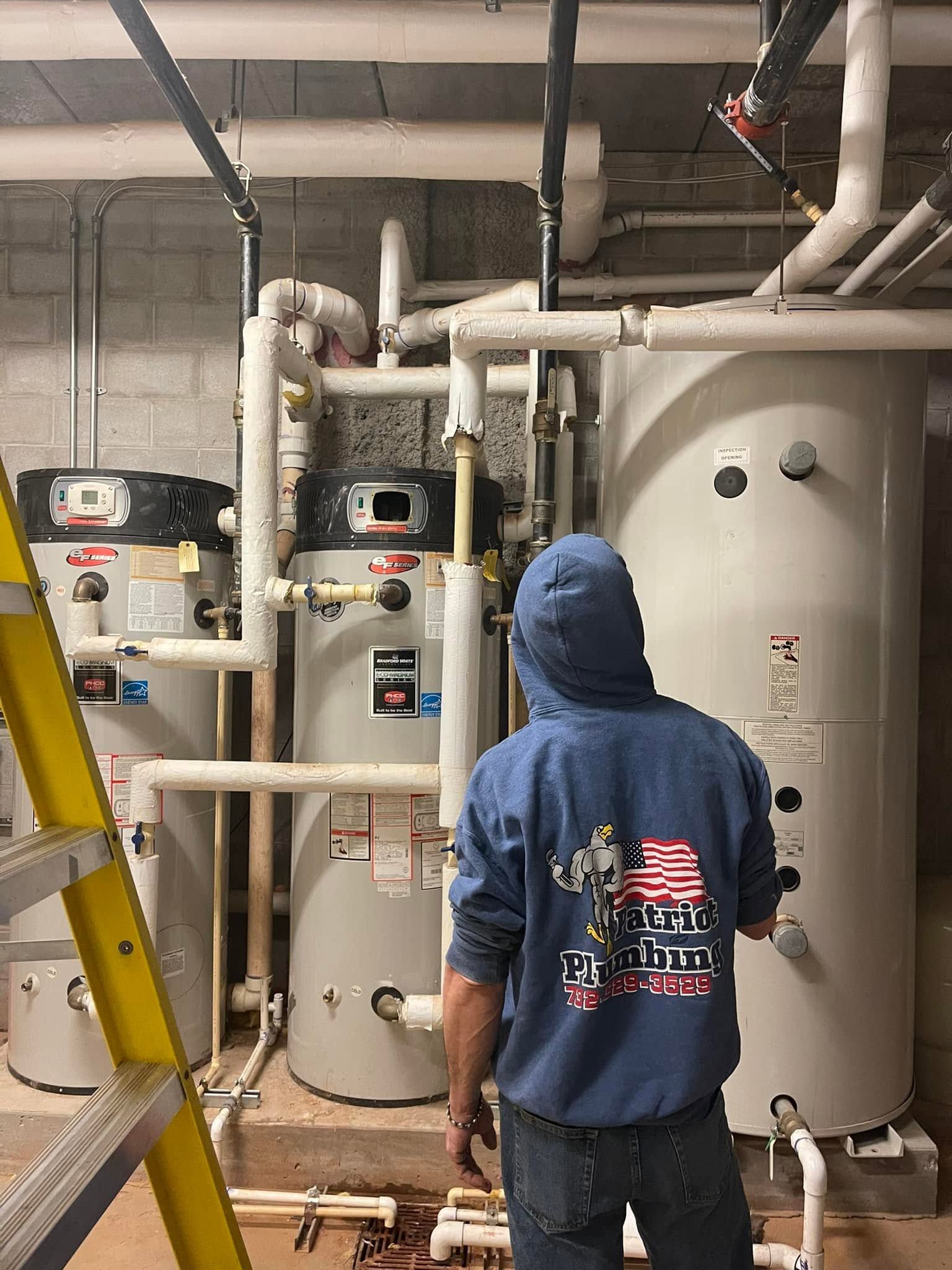 Worker in blue hoodie inspecting industrial boilers and piping in a mechanical room.