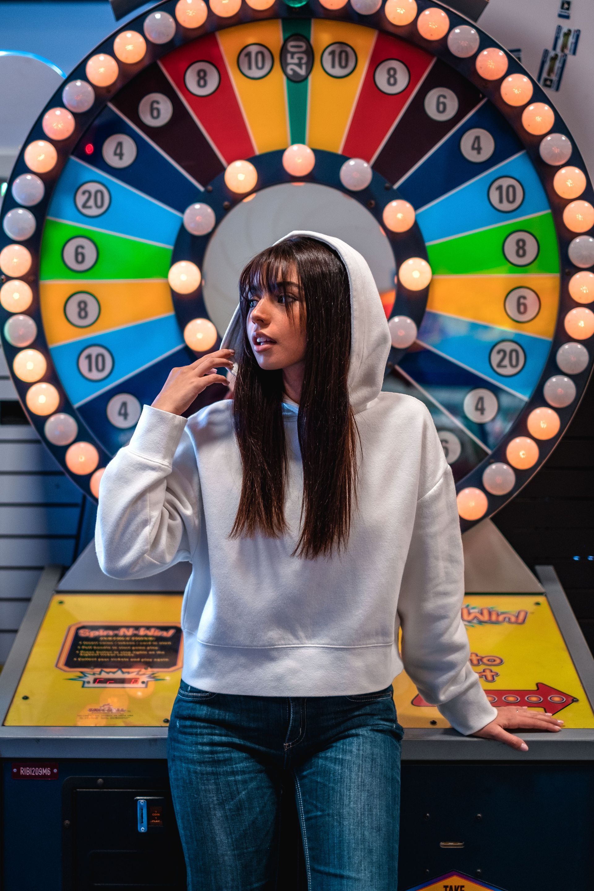 Woman in gray hoodie standing at a colorful carnival prize wheel, holding a microphone.