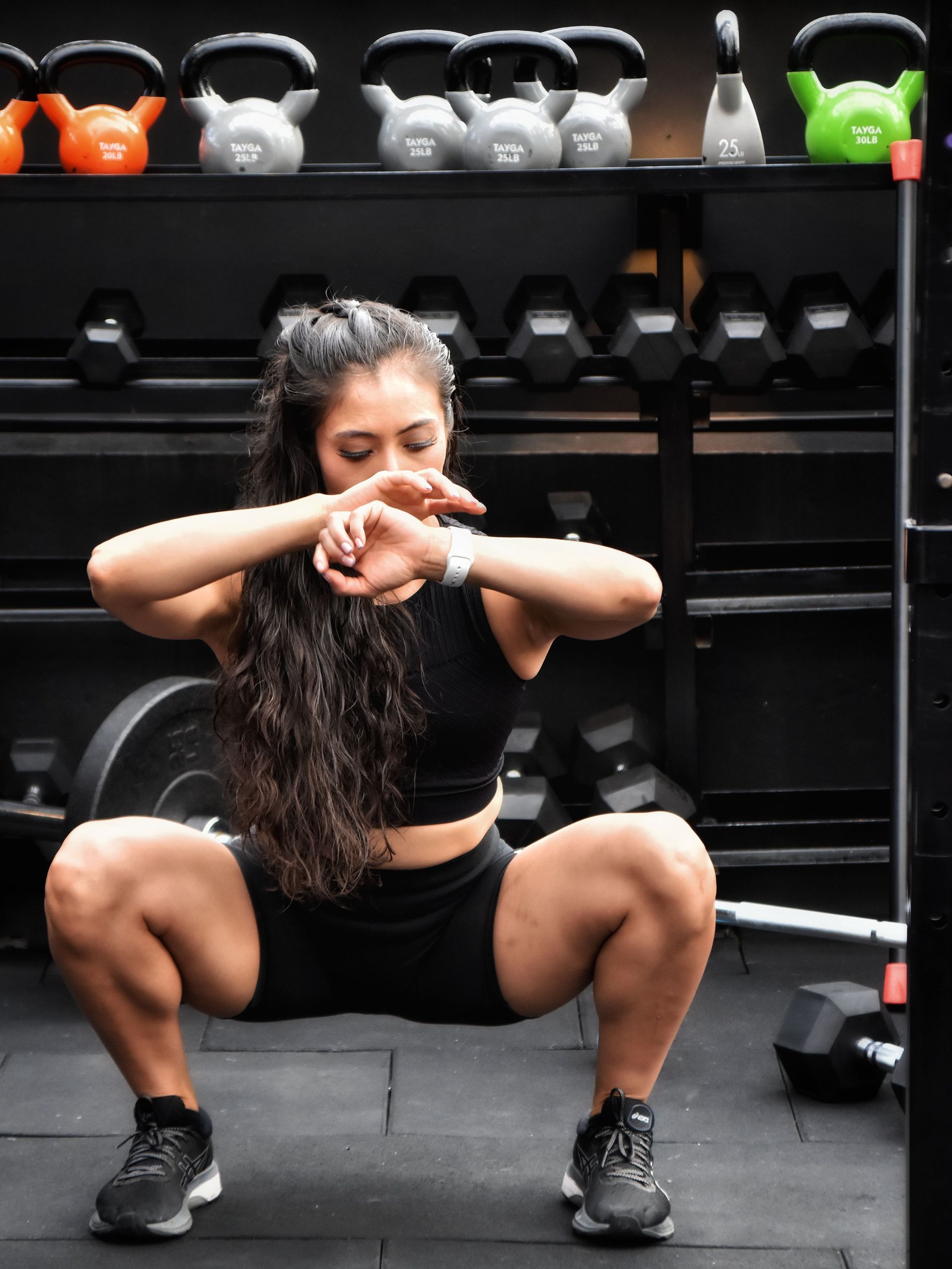 Person doing a deep squat workout in a gym, with kettlebells and dumbbells on shelves behind them.