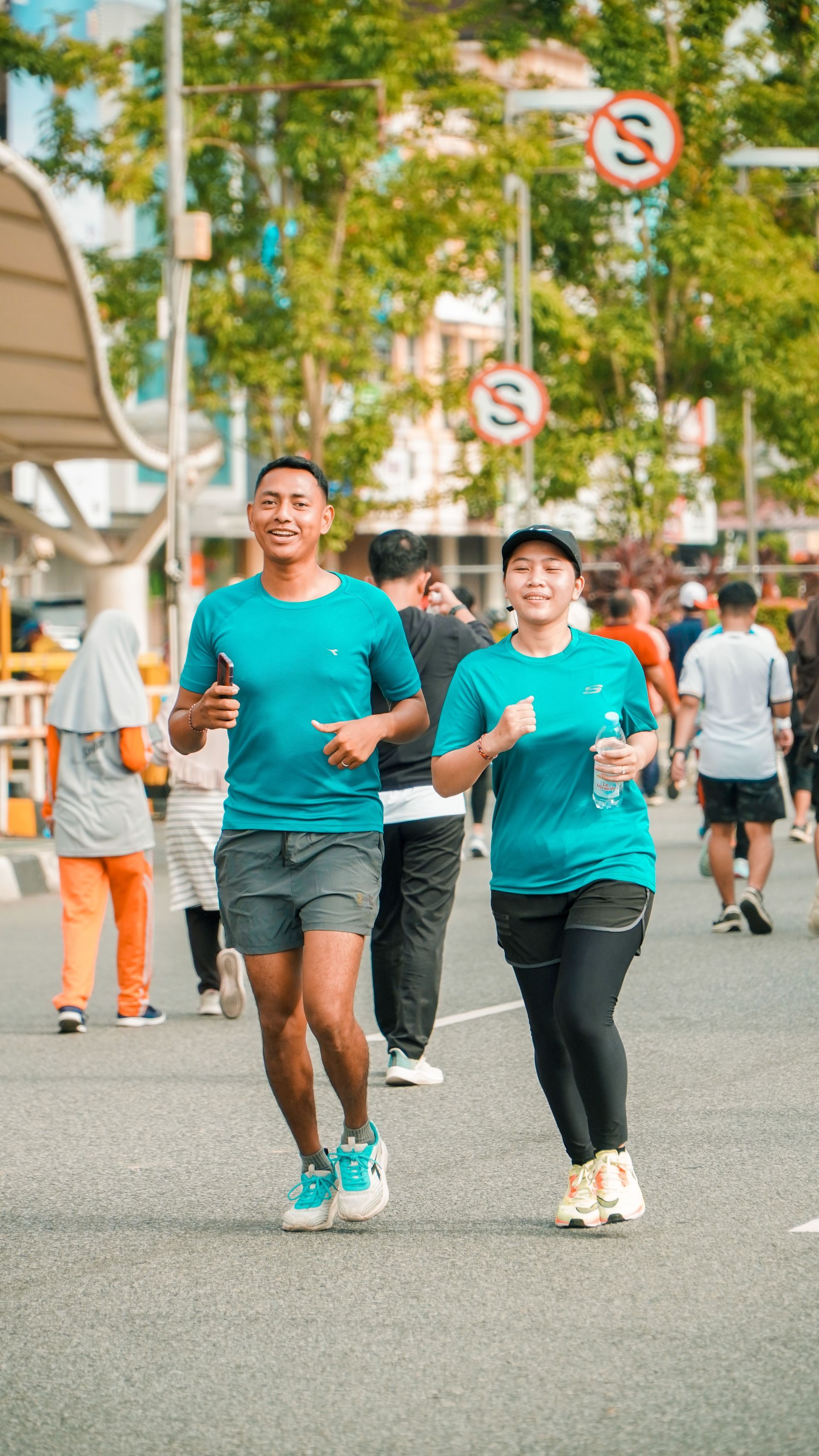 Two runners in teal shirts jogging on a city street with other people in the background