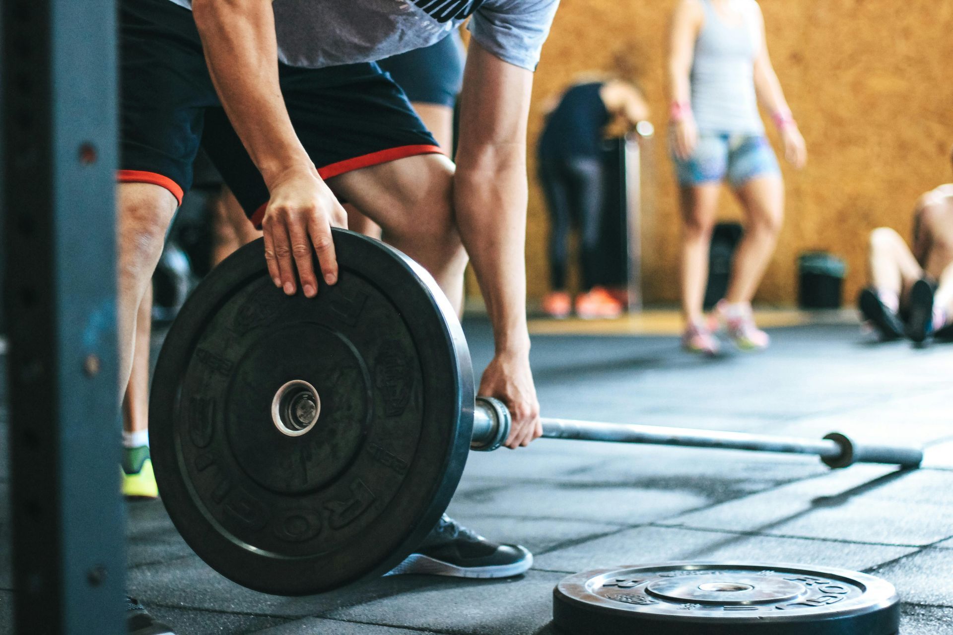 Person loading a barbell plate in a gym, with other athletes exercising in the background.