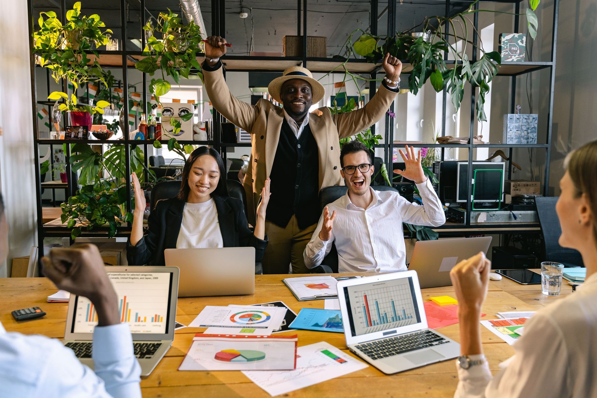 Team in a bright office celebrates around a table with laptops, charts, and raised arms