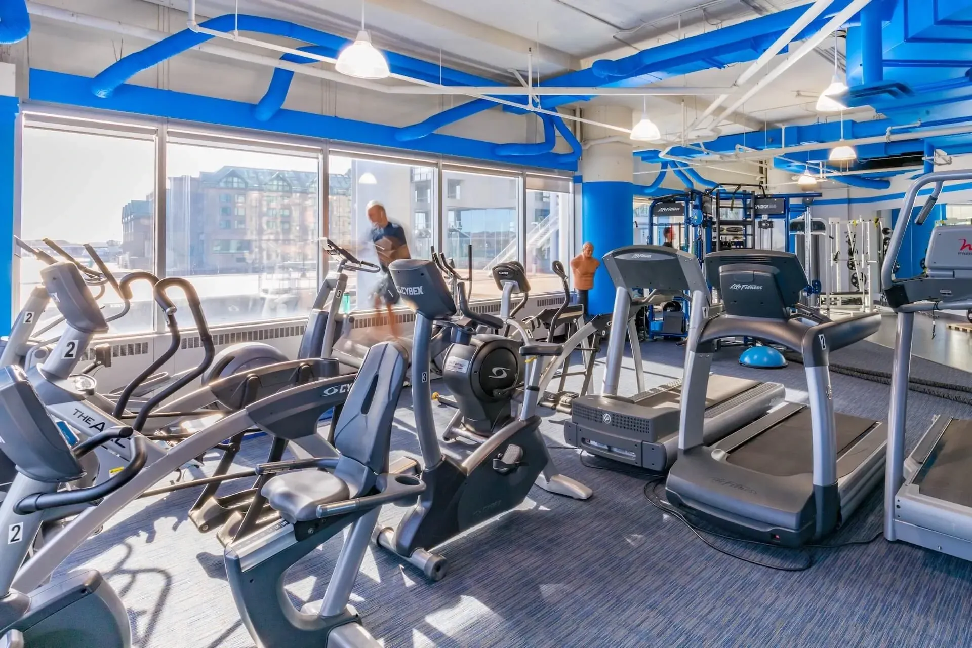 Bright gym with treadmills and exercise bikes under blue ceiling beams, overlooking a city view