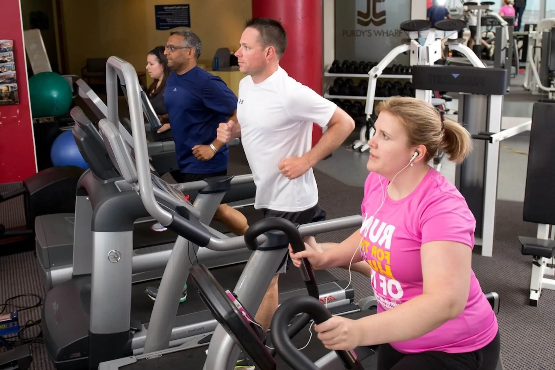 People exercising on cardio machines in a gym, including a woman in a pink shirt in the foreground.