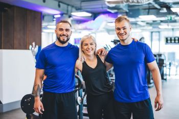 Three people in blue workout shirts smiling in a gym, standing arm in arm.
