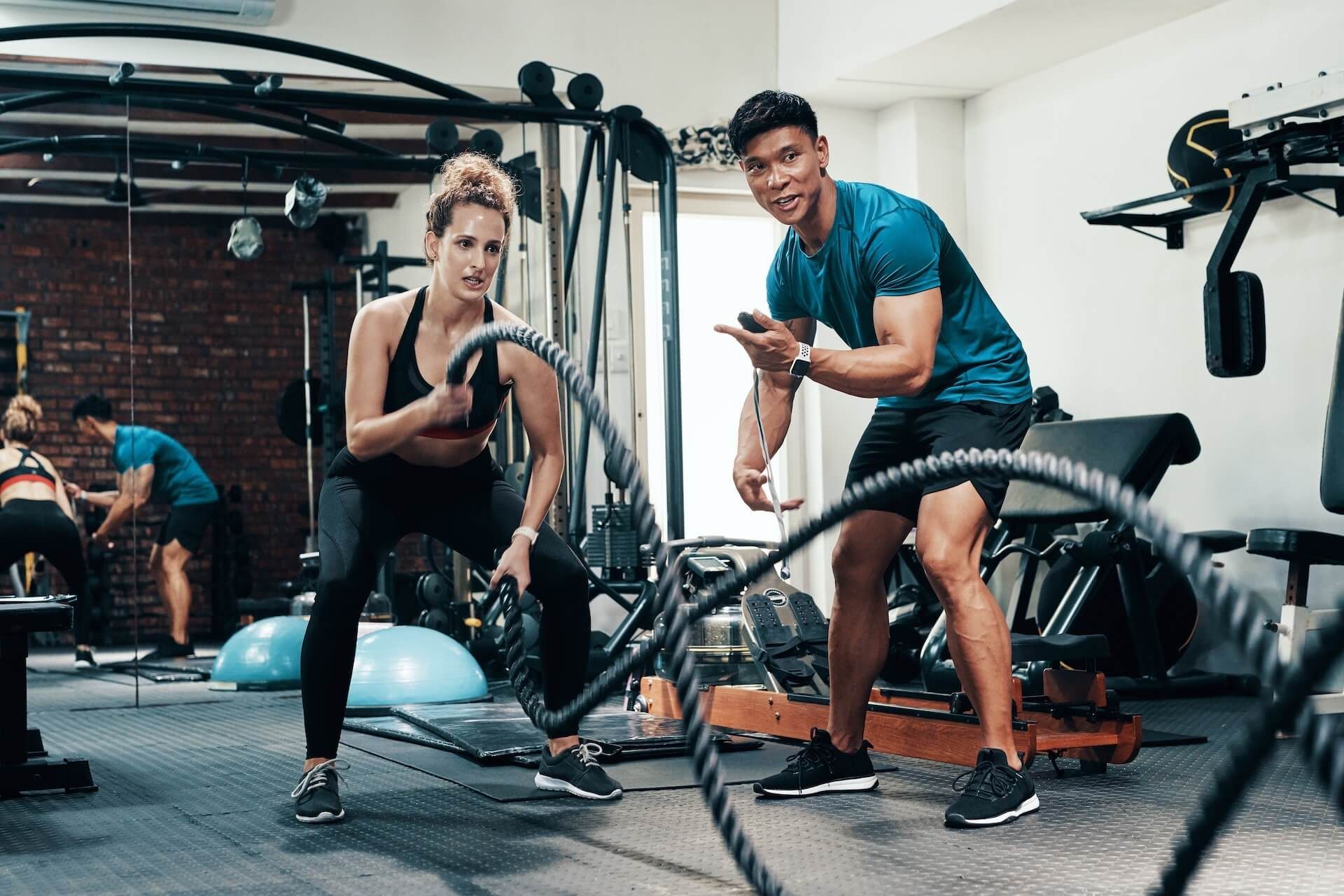 Two people exercising with battle ropes in a gym, one smiling and one focused.