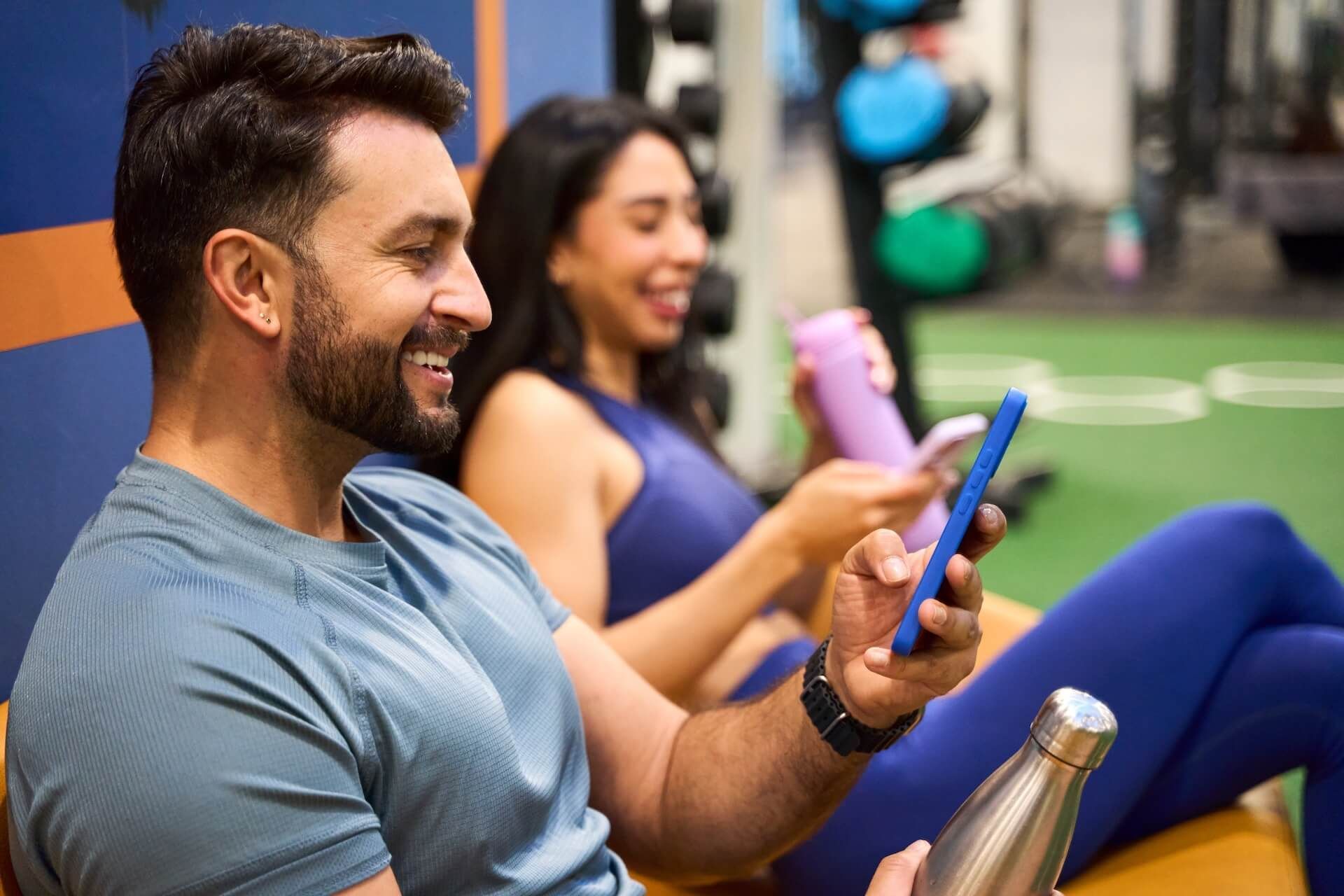 Two people relaxing on gym mats, smiling while using smartphones with water bottles nearby.